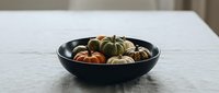 Colorful mini pumpkins and gourds arranged in black ceramic bowl on white marble surface for autumn seasonal home decor and fall harvest celebration.