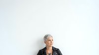 Mature woman with short gray hair wearing black leather jacket against white wall background.