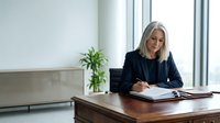 Mature woman with shoulder-length blonde hair, Caucasian ethnicity, wearing dark blazer, writing in notebook at wooden desk in modern office with plant and laptop. Business professional.