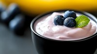Fresh blueberries and mint leaf garnish on creamy pink yogurt dessert in black ceramic bowl, selective focus on berries against blurred background.