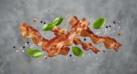 Crispy bacon strips flying with fresh basil leaves and peppercorns on gray background. Food ingredients in motion for cooking and culinary projects.