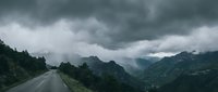 Asphalt road stretching through misty mountain valley with dense forest and dramatic storm clouds overhead, suitable for travel and nature backgrounds.