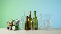 Assorted recyclable materials including glass bottles, aluminum cans and plastic containers arranged on wooden surface against gradient background for sorting.