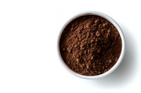Ground cocoa powder in white ceramic bowl on white background, overhead view showing rich dark brown color and fine texture. Isolated food ingredient.