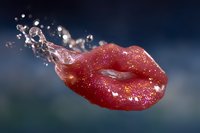 Glossy red lips with glitter and water droplets splashing in dynamic motion against dark blue background, creating dramatic cosmetic beauty effect.