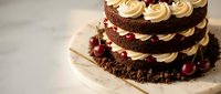 Decadent chocolate layer cake with cream frosting and cherry garnish on rustic wooden surface for bakery menu dessert photography.