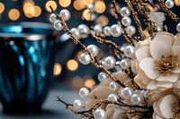 Elegant pearl beaded branches with cream fabric flower against blurred bokeh lights and blue glass vase, creating luxurious holiday or wedding decor atmosphere.