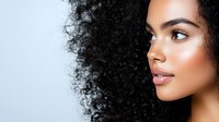 Young African American woman with curly hair and natural makeup looking sideways in profile view against light background for beauty and skincare concepts.