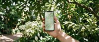 Hand holding smartphone with Follow Us screen in lush green tropical garden, social media concept for eco brands and nature businesses.