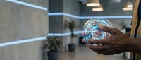 Hand holding transparent globe sphere with digital network connections inside modern office environment with blurred background and plants.