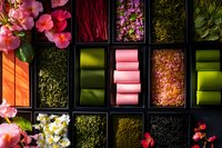 Colorful assortment of tea ingredients in black compartment box with dried flowers, herbs, and rolled pink tea leaves, styled for food photography and packaging design.