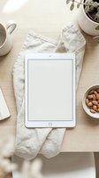 White tablet with blank screen on linen kitchen towel surrounded by coffee cup, almonds and plant creating cozy workspace mockup for digital content display.