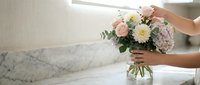 Woman arranging fresh pink and white flower bouquet in glass vase on marble countertop in bright modern kitchen interior for home decoration.
