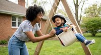 African American mother and toddler son playing on wooden swing in backyard garden. Happy family outdoor activity with child wearing safety helmet.