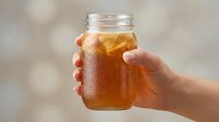 Caucasian hand holding glass jar filled with cold iced tea or cola beverage against blurred outdoor background, refreshment drink concept.