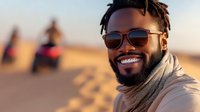 Young African American man with dreadlocks and sunglasses smiling broadly at sunset, wearing beige scarf, warm golden lighting creates positive emotional atmosphere.