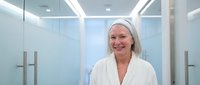 Happy mature Caucasian woman in white bathrobe and headband smiling in modern spa bathroom with glass shower doors and contemporary lighting fixtures.