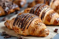 Fresh baked croissants drizzled with dark chocolate sauce and sprinkled with chocolate shavings on parchment paper, close-up view of French pastries.