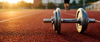 Pair of dumbbells resting on outdoor running track at golden hour, with blurred jogger in background. Fitness and training concept.