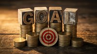 Wooden letter blocks spelling GOAL on stacks of golden coins with red target bullseye, representing financial objectives and business success planning.