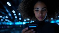 Young African American woman with natural curly hair looking at mobile phone screen in dark environment with blue bokeh lights creating modern urban nighttime atmosphere.