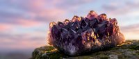 Large purple amethyst crystal cluster on moss-covered rock against dramatic sunset sky with pink and purple clouds for healing and spiritual wellness concepts.