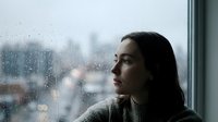 Young woman gazing thoughtfully through rain-covered window with city lights blurred in background, conveying solitude and contemplation indoors.