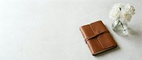 Brown leather journal notebook with elastic band closure on neutral background with white flowers for writing planning and organization.