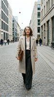Professional middle-aged Caucasian businesswoman in beige coat walking confidently through modern urban street with cobblestones and contemporary architecture.