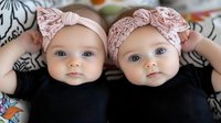 Twin baby girls wearing matching pink lace headbands and black outfits lie side by side, gazing directly at camera with bright blue eyes and innocent expressions.