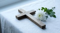 Wooden cross with white chrysanthemum flower on soft linen fabric creating peaceful memorial or religious ceremony atmosphere for spiritual reflection.