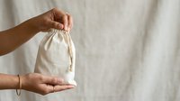 Hands holding small natural cotton drawstring pouch against neutral background for product packaging, storage, and eco-friendly lifestyle concepts.