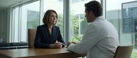 Professional businesswoman and businessman having serious discussion in modern office conference room with large windows and city view.
