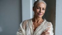 Elegant mature woman with silver hair wearing delicate lace wrap, smiling confidently by window with soft natural light. Perfect for wellness, beauty, or aging gracefully concepts.