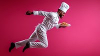 Young male chef in white uniform and hat levitating while holding pizza against vibrant pink background, dynamic action pose capturing culinary movement.