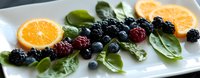 Fresh mixed berries with orange slices and green spinach leaves arranged on white rectangular plate, colorful healthy food composition for breakfast or dessert.