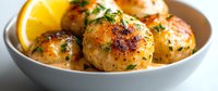 Juicy chicken meatballs in creamy garlic sauce garnished with fresh herbs and lemon slice, served in white ceramic bowl on light background.