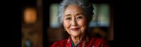 Elegant senior Asian woman with gray hair wearing red traditional clothing smiling warmly in soft indoor lighting for lifestyle and cultural content.