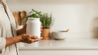Unrecognizable person holding blank white jar in modern kitchen with potted herbs and neutral background for product mockup.