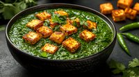 Palak paneer curry with crispy golden cheese cubes floating in creamy pureed spinach sauce, garnished with fresh herbs and green chilies on dark background.