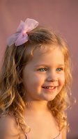 Adorable young girl with curly blonde hair and pink bow smiling warmly in soft golden light against pastel background for children photography.