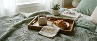 Cozy breakfast in bed with croissant, hot coffee, and Stay Home napkin on wooden tray, green knit blanket, morning relaxation concept.