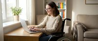 Woman with dark hair and glasses, appearing to be of Caucasian ethnicity, working on laptop while seated in wheelchair at wooden desk in bright home office with bookshelf and couch nearby.
