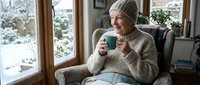Senior Caucasian woman in cozy knit sweater and beanie enjoying hot beverage by window during winter, relaxing at home with warm drink and snowy garden view.