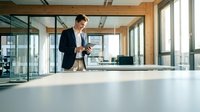Caucasian male professional in dark business suit standing at modern white conference table in bright office space with wooden beams and floor-to-ceiling windows, suitable for corporate, business, and workplace concepts.