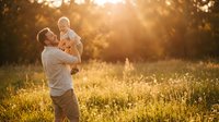Young Caucasian father lifting happy baby in sunny meadow at golden hour, joyful family bonding moment outdoors in warm summer light.