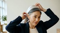 Asian woman in professional black blazer and cream sweater adjusting gray headphones around her neck in bright office space with window and plant, suitable for corporate, technology, and business communications.
