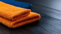 Folded orange and blue microfiber cleaning cloths on dark wooden surface, close-up view showing texture detail and fabric layers for household cleaning concept.