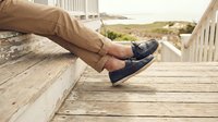Man's legs in beige chinos and navy boat shoes resting on weathered wooden beach house steps with ocean view in background.