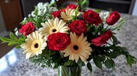 Fresh floral arrangement with vibrant red roses and cream gerbera daisies in glass vase, accented with white baby's breath and green foliage on marble counter.
